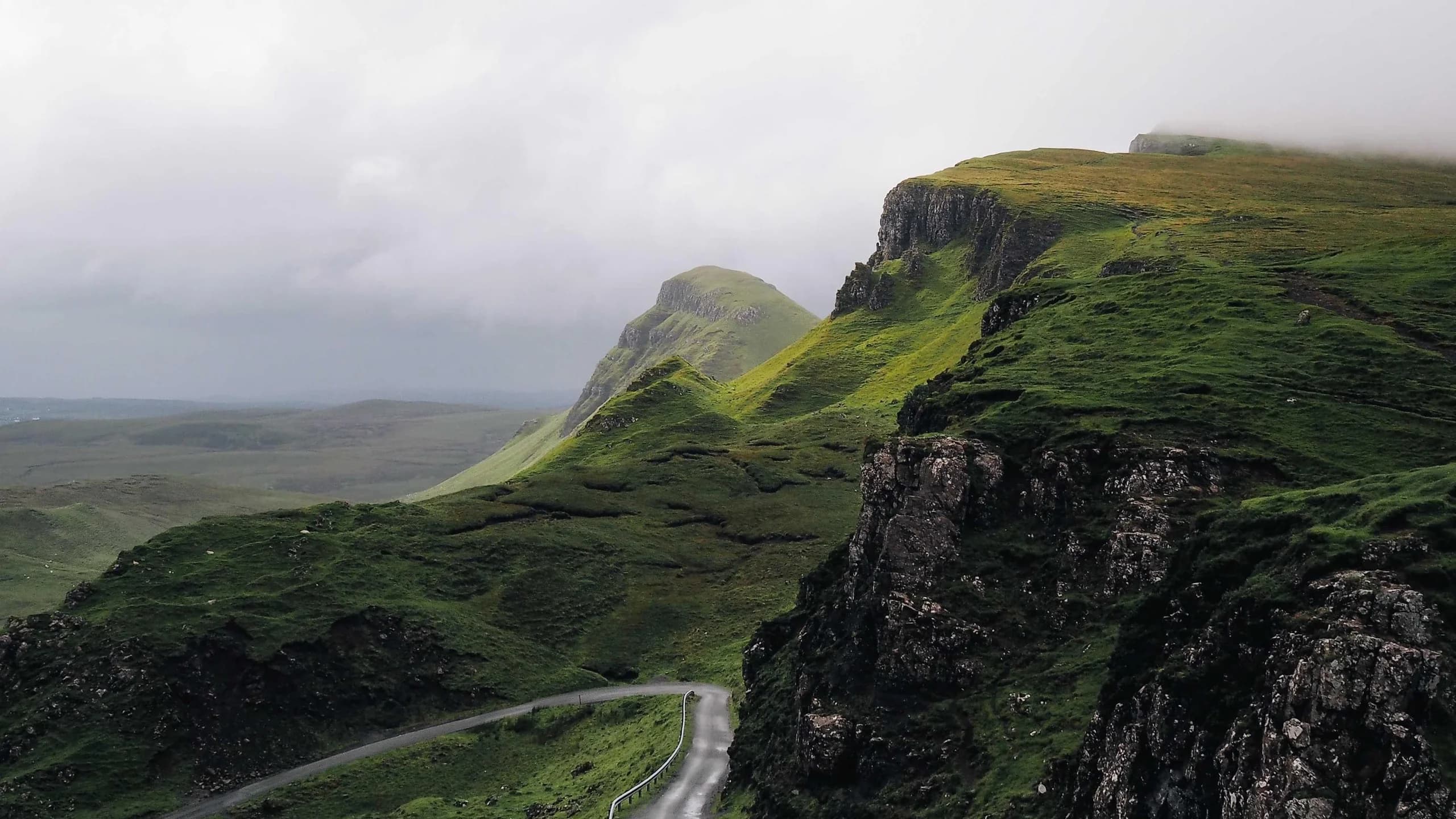 Crête verte et falaises plongeant vers une route, sous un ciel d’orage